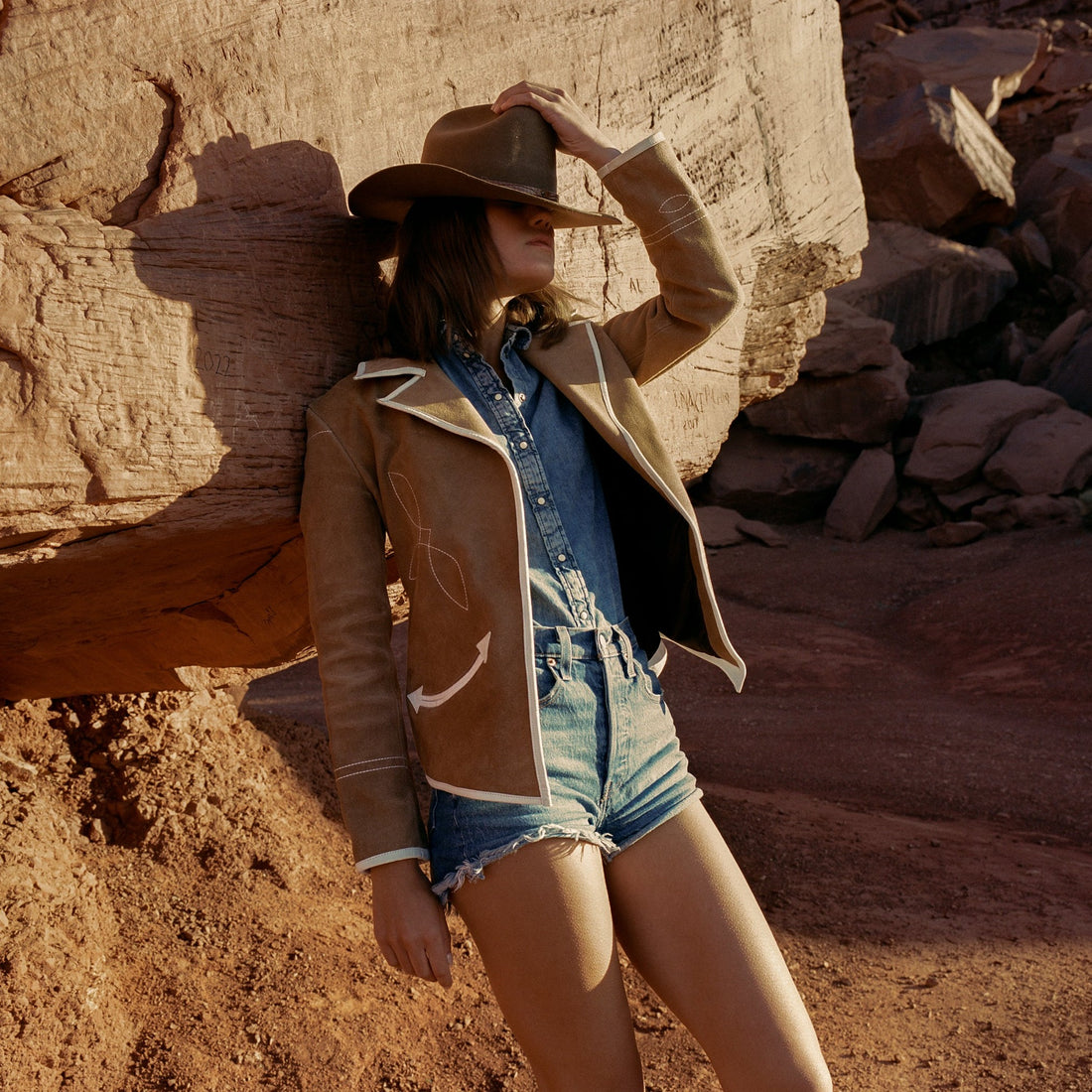 A woman wearing a caramel suede blazer with white leather piping, paired with a denim shirt and shorts, standing near rocky terrain.