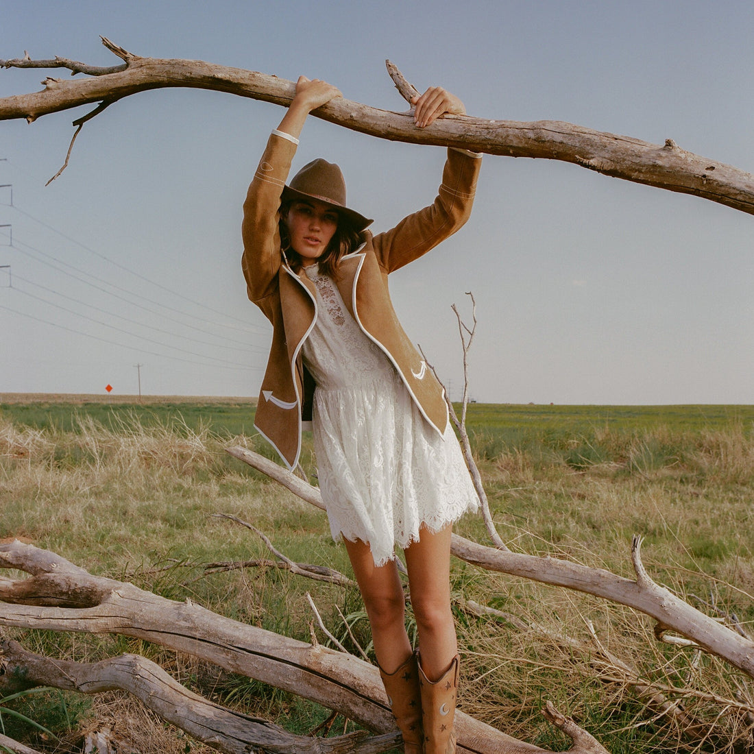 A woman standing on a tree trunk, wearing a brown carmel suede blazer with white leather piping and details in a white dress