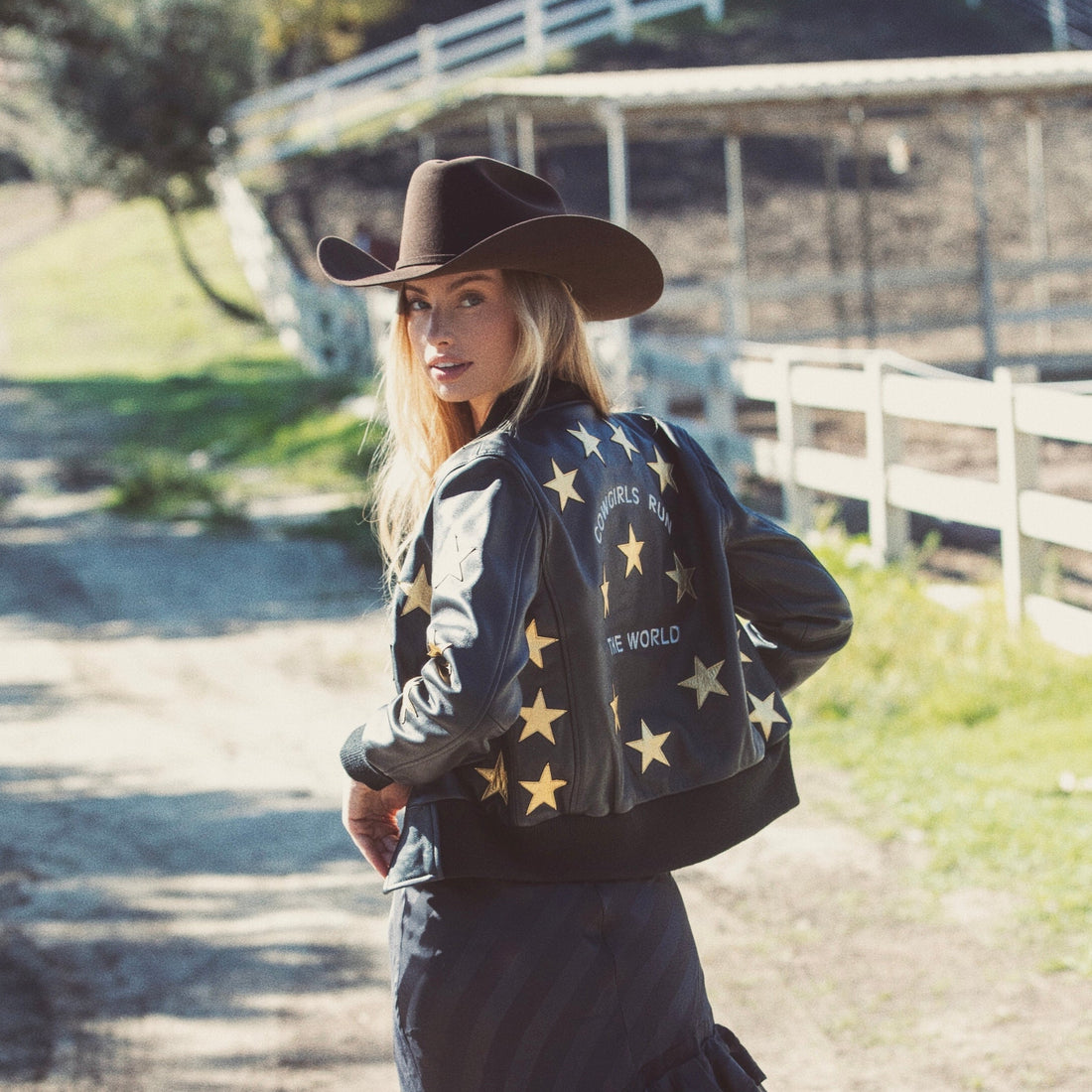A woman wearing a black bomber jacket with metallic gold stars and text 'Cowgirls Run the World' on the back.