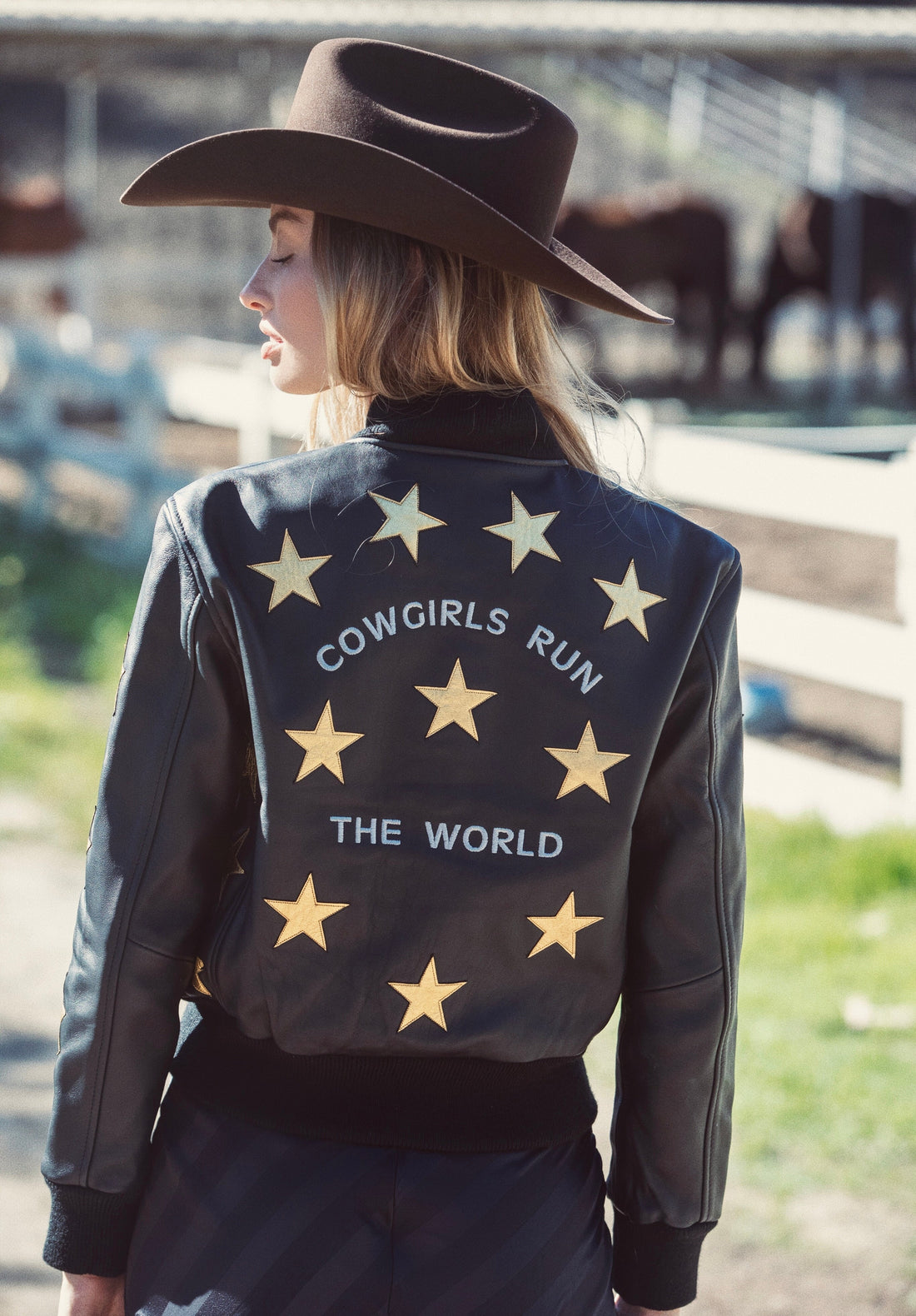 A woman wearing a black bomber jacket with metallic gold stars and text 'Cowgirls Run the World' on the back.