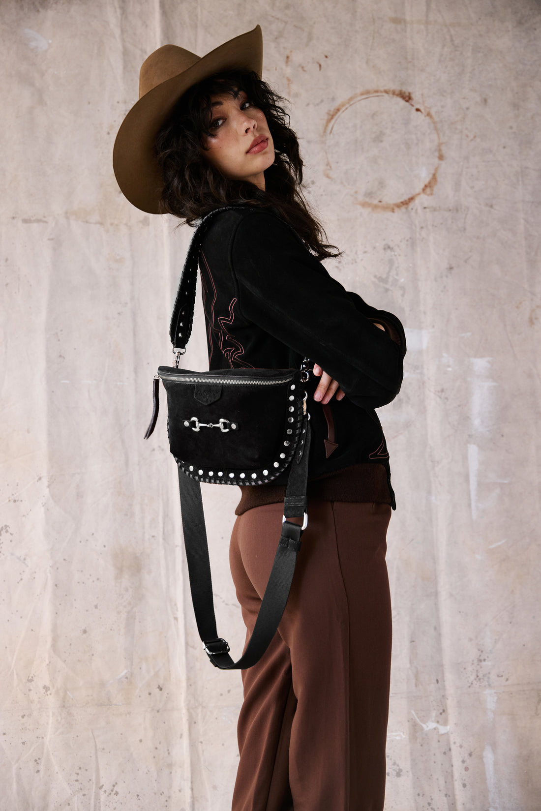 A woman in a cowboy hat wearing a black suede bum bag with silver hardware and two removable straps, one over the shoulder and one across the body. Features a horse bit on the front