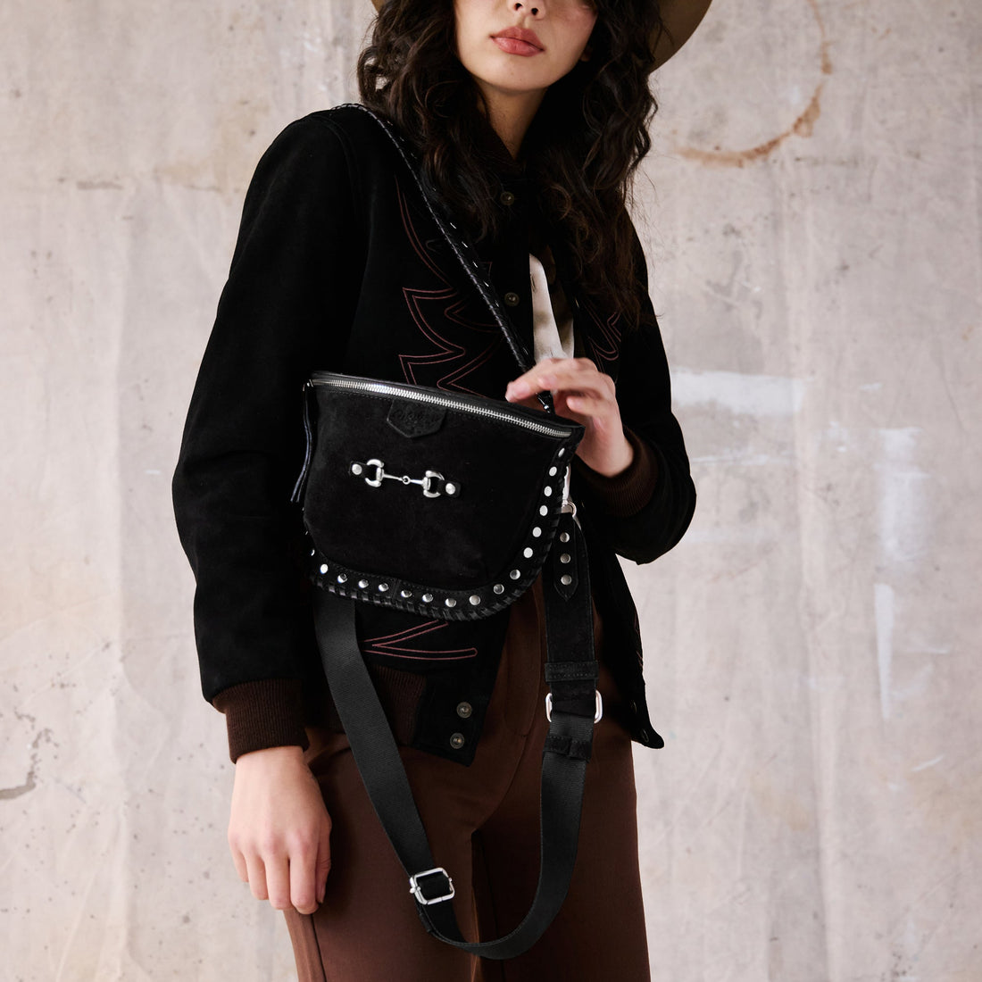 A woman wearing a black suede bum bag with silver hardware and two removable straps, one over the shoulder and one across the body. Features a horse bit on the front