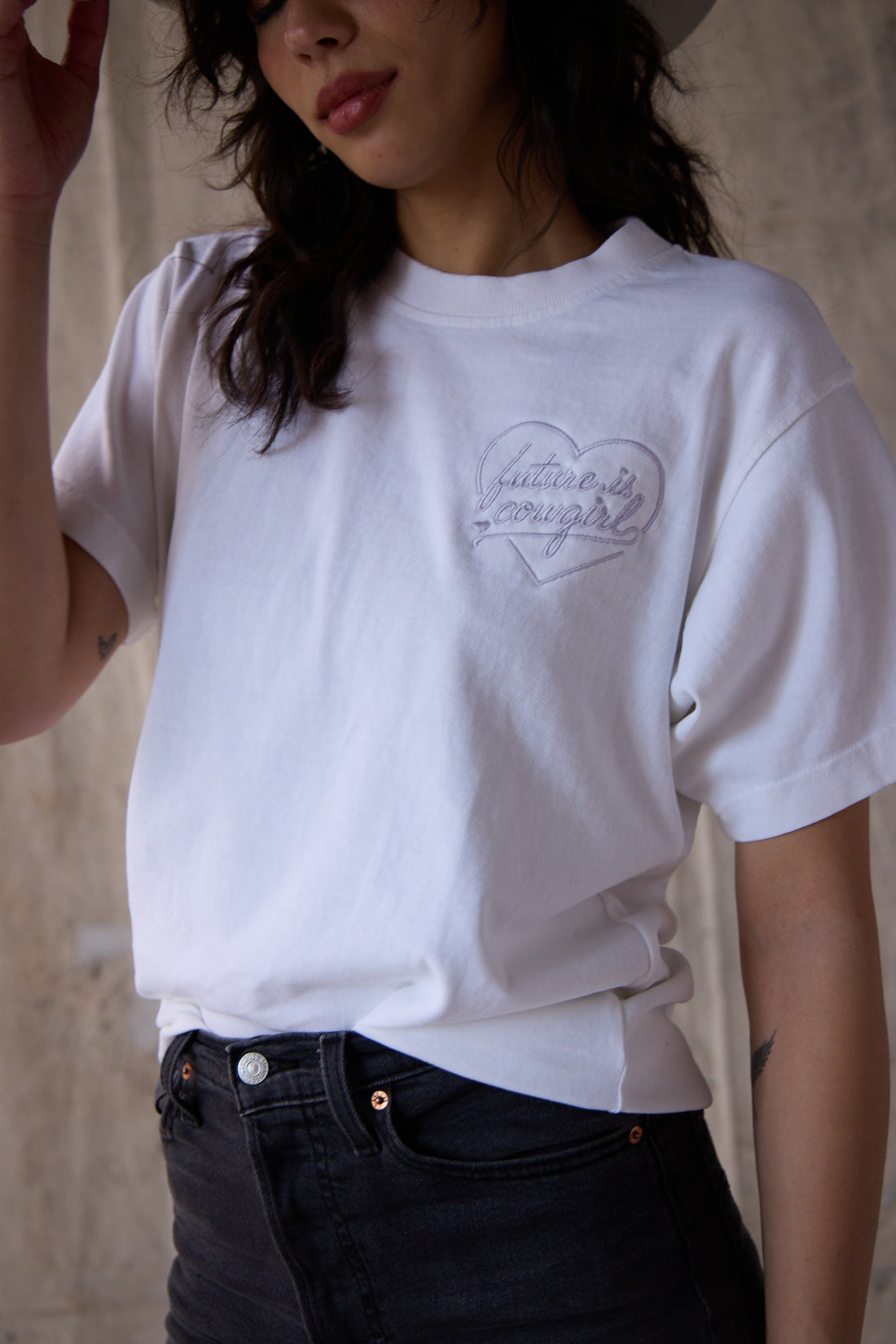 A woman wearing a white t shirt with an embroidered heart future is cowgirl logo