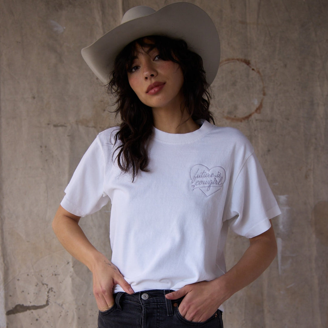 A woman wearing a white t shirt with an embroidered heart future is cowgirl logo