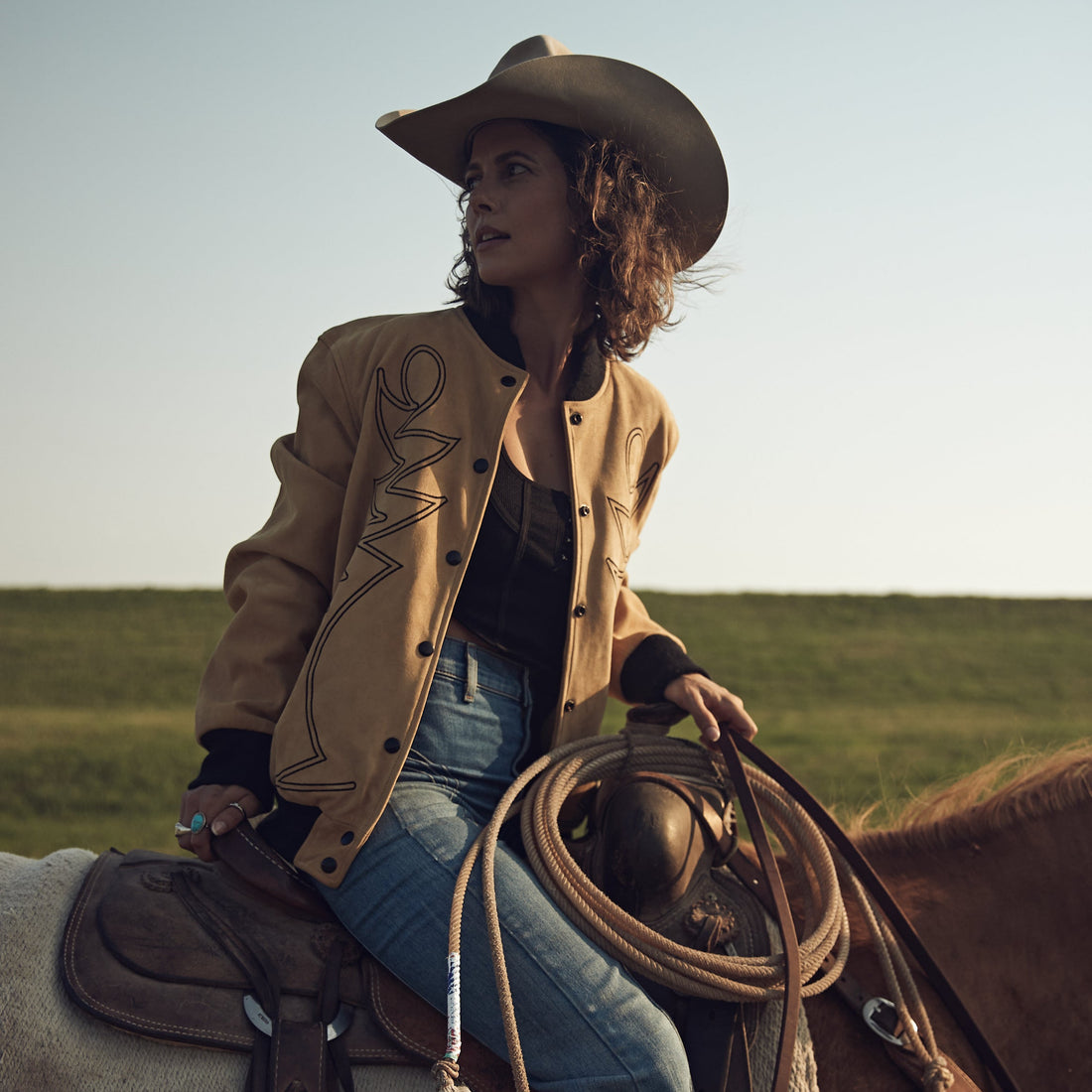 A woman riding  a horse in a field wearing a beige embroidered bomber jacket with black western embroidery, paired with blue jeans and a black top.