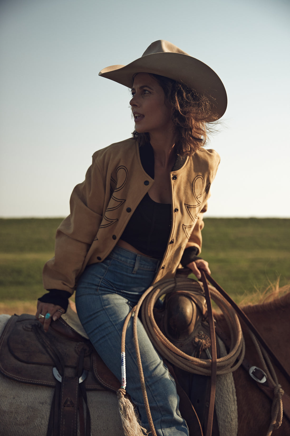 A woman riding a horse in a field wearing a beige embroidered bomber jacket with black western embroidery, paired with blue jeans and a black top.
