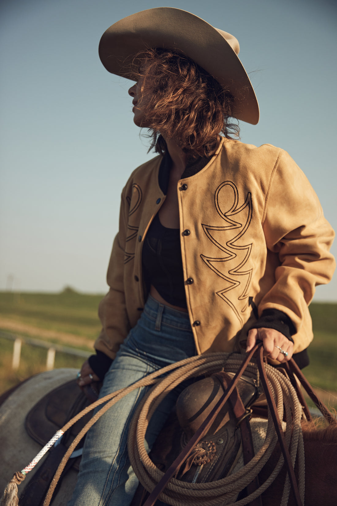 A woman riding a horse in a field wearing a beige embroidered bomber jacket with black western embroidery, paired with blue jeans and a black top.