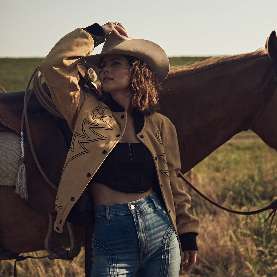 A woman standing by a horse in a field wearing a beige embroidered bomber jacket with black western embroidery, paired with blue jeans and a black top.