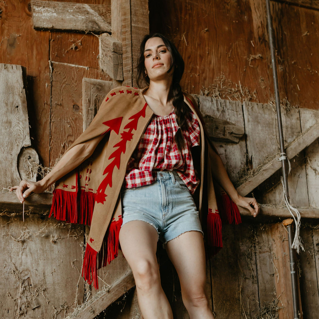 A woman wearing a beige and red suede cape with fringe, paired with a plaid shirt and denim shorts, standing in front of a rustic wooden backdrop.