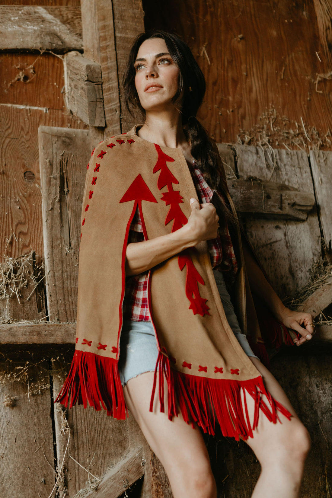A woman wearing a beige and red suede cape with fringe, paired with a plaid shirt and denim shorts, standing in front of a rustic wooden backdrop.