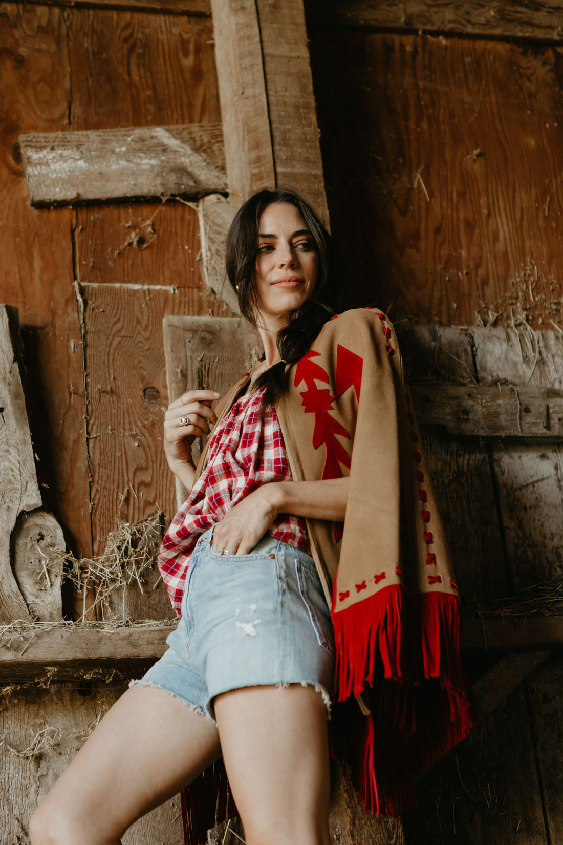 A woman wearing a beige and red suede cape with fringe, paired with a plaid shirt and denim shorts, standing in front of a rustic wooden backdrop.