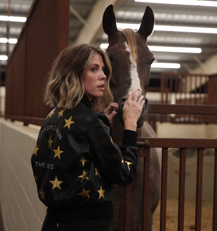 A woman standing next to a horse, wearing a black bomber jacket with metallic gold stars and text 'Cowgirls Run the World' on the back.