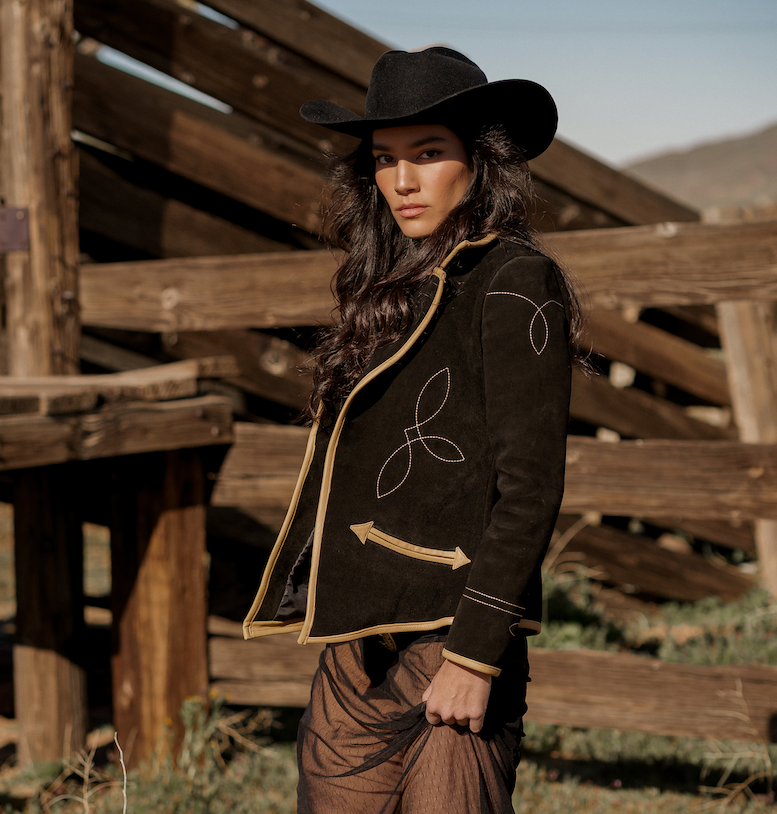 A woman wearing a black suede blazer with tan leather details, paired with a hat and boots, standing in front of a wooden fence.