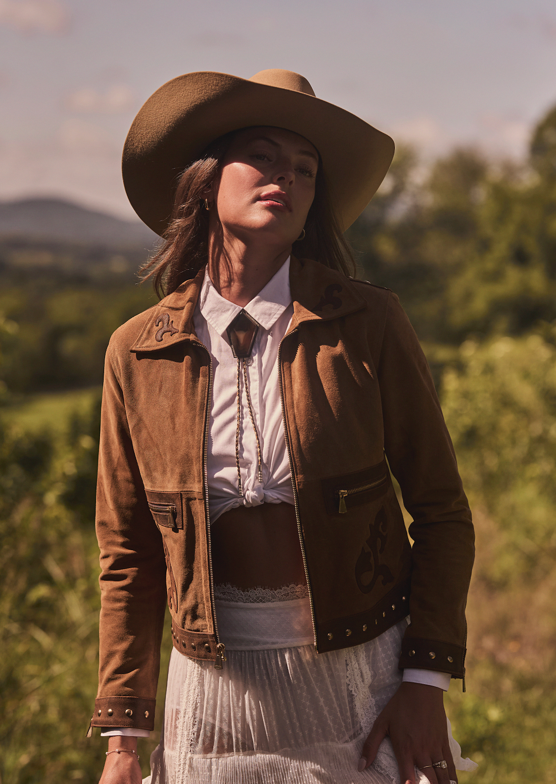 Woman wearing a cowboy hat and brown embroidered jacket by Western & Co in a natural outdoor setting