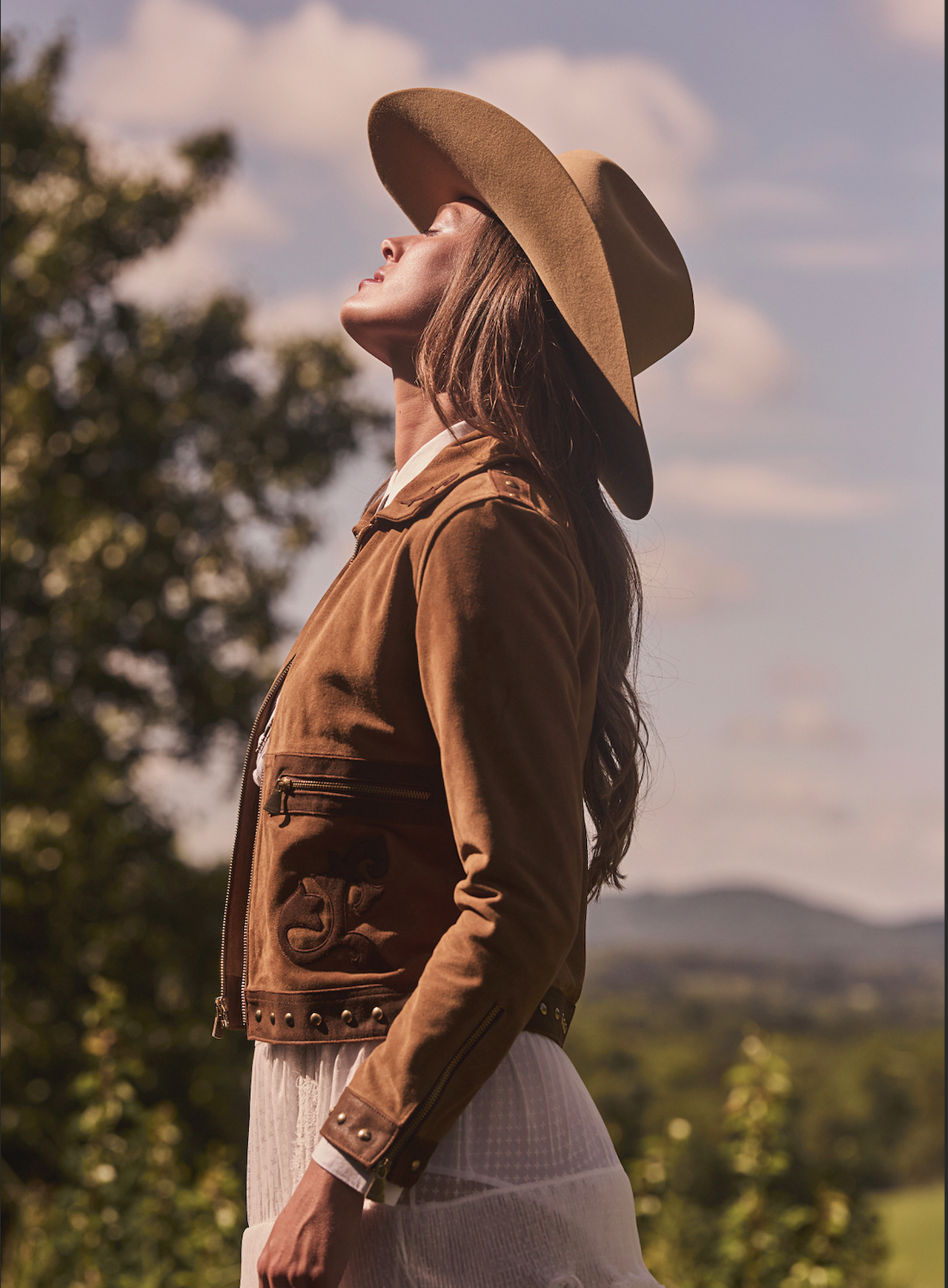 Woman wearing a cowboy hat and brown embroidered jacket by Western & Co standing outside with trees in the background