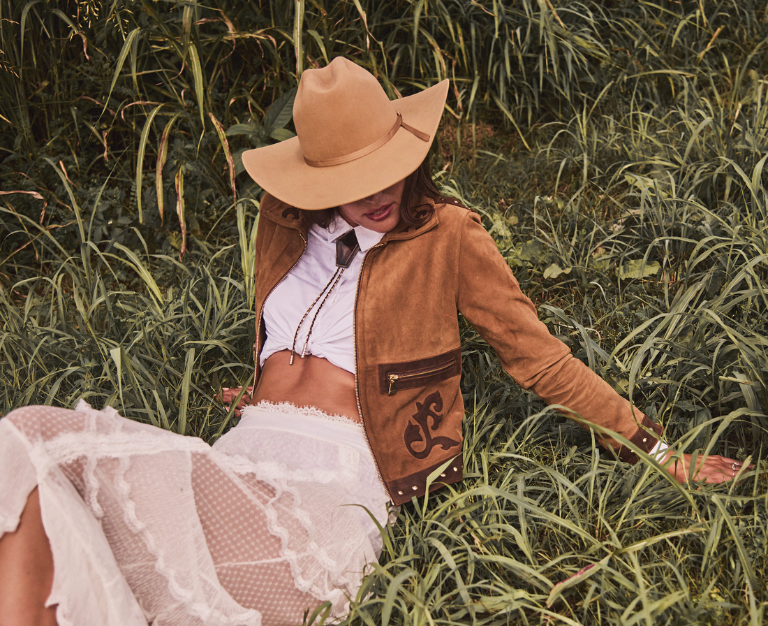 Woman wearing a cowboy hat and brown embroidered jacket by Western & Co in a grassy field