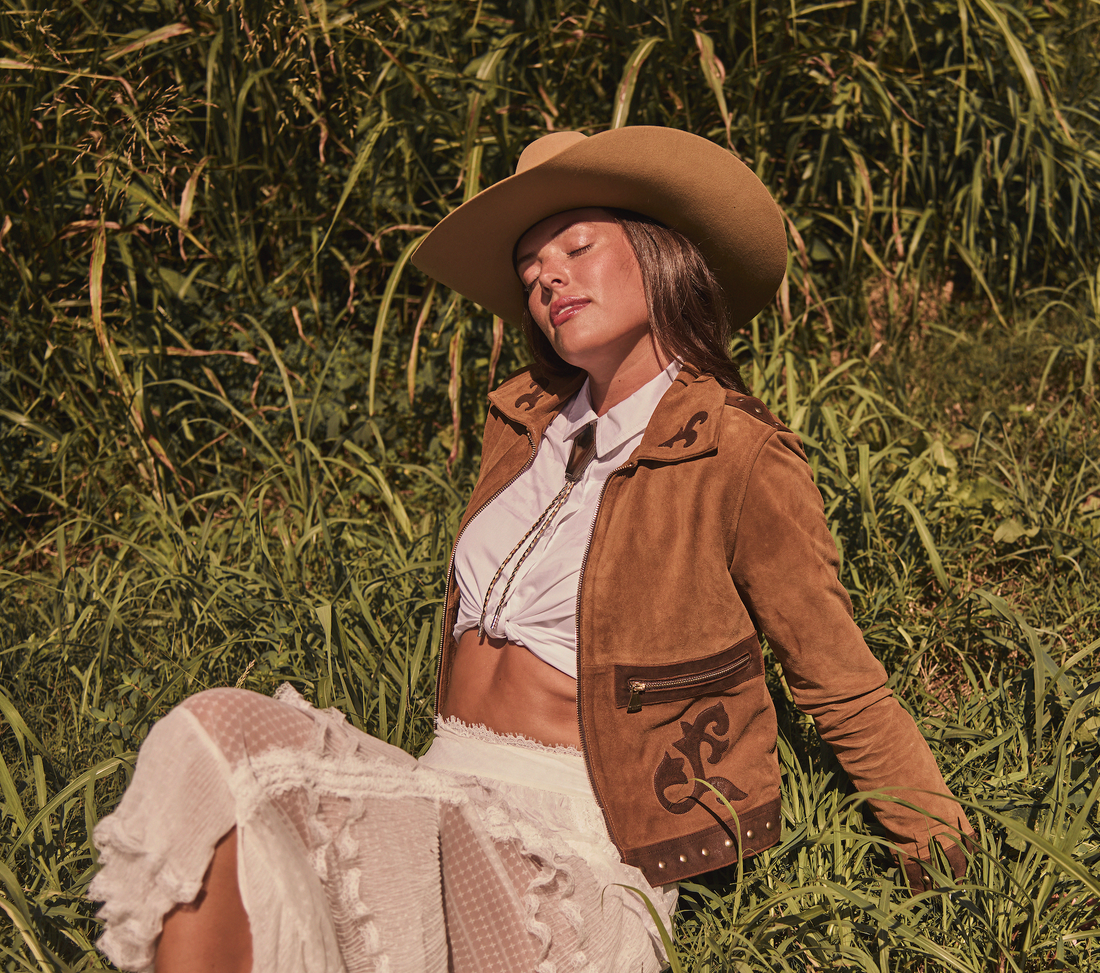 Woman wearing a cowboy hat and brown embroidered jacket by Western & Co in a grassy field