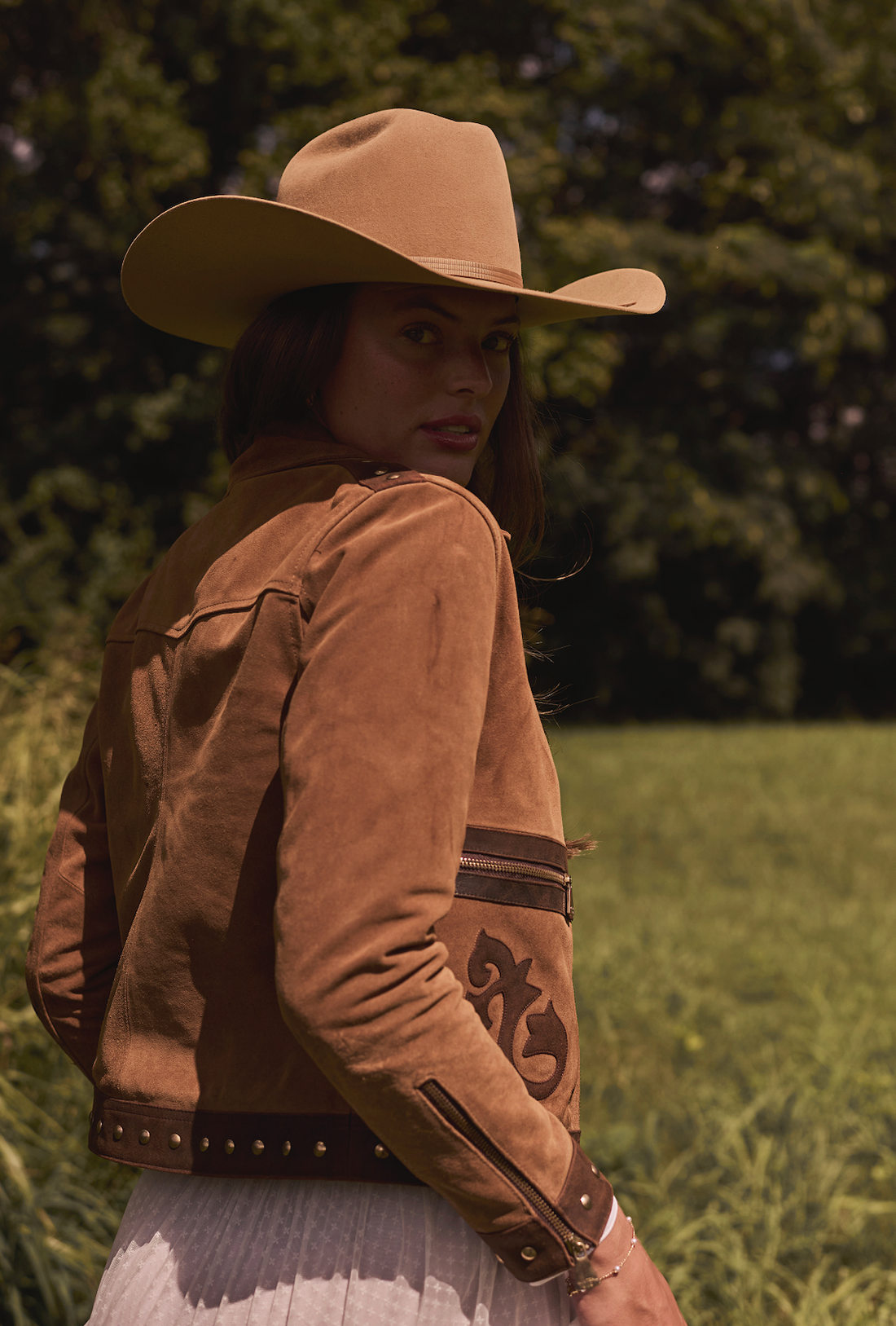 Woman wearing a cowboy hat and brown embroidered jacket by Western & Co in a grassy field