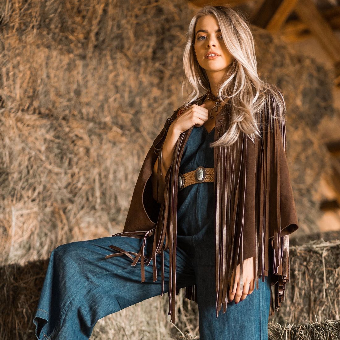 A woman wearing a brown suede cape with long metallic bronze fringe standing in front of a hayloft.