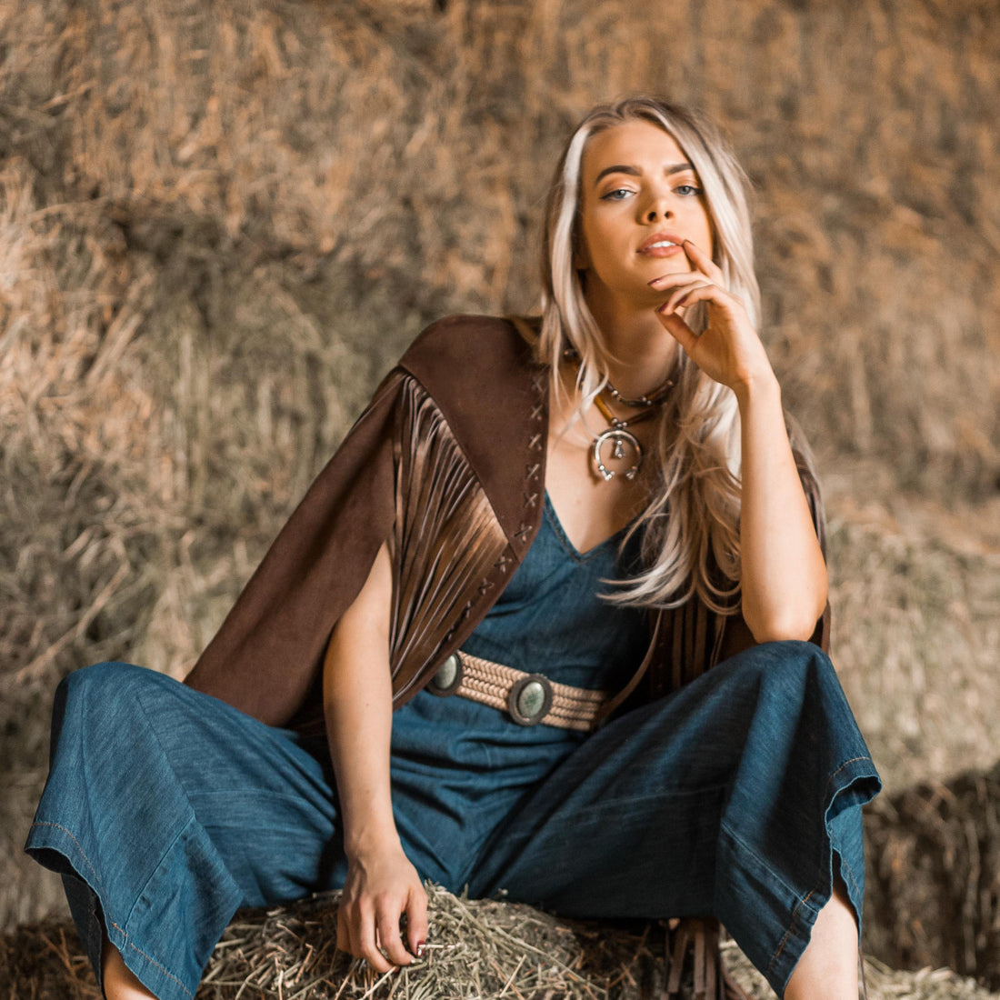 A woman wearing a brown suede cape with long metallic bronze fringe sitting on a hay bale