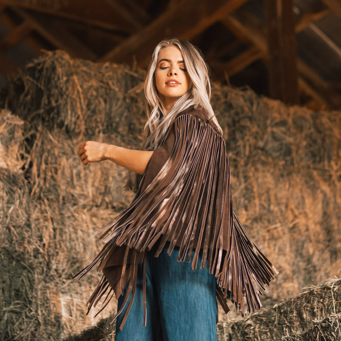 A woman wearing a brown suede cape with long metallic bronze fringe standing in front of a hayloft.