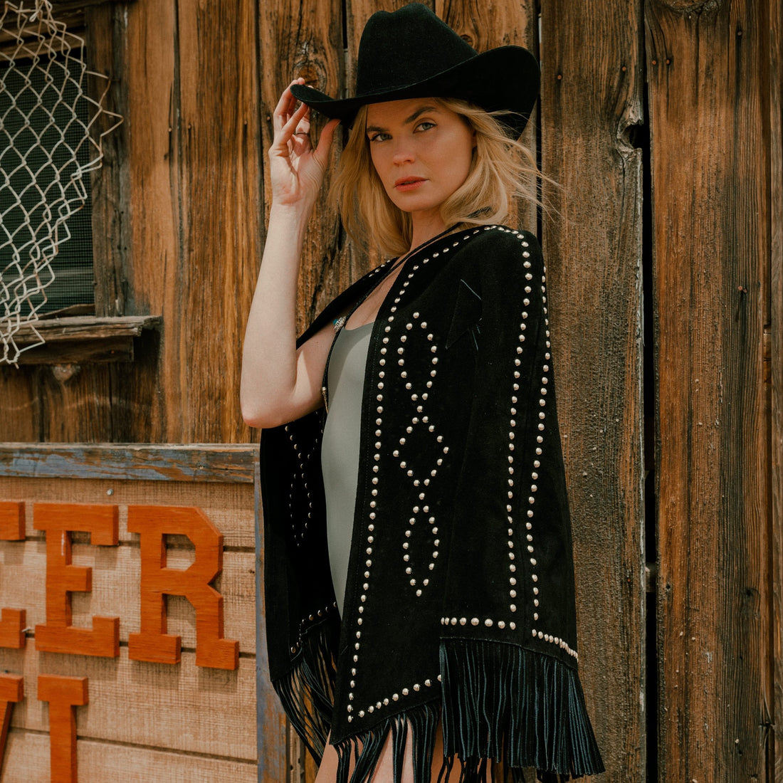 A woman wearing a black suede cape with silver stud details and fringe, paired with a cowboy hat