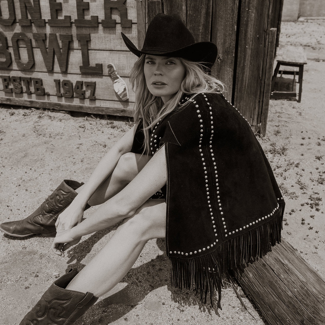 A woman wearing a black suede cape with silver stud details and fringe, paired with a cowboy hat