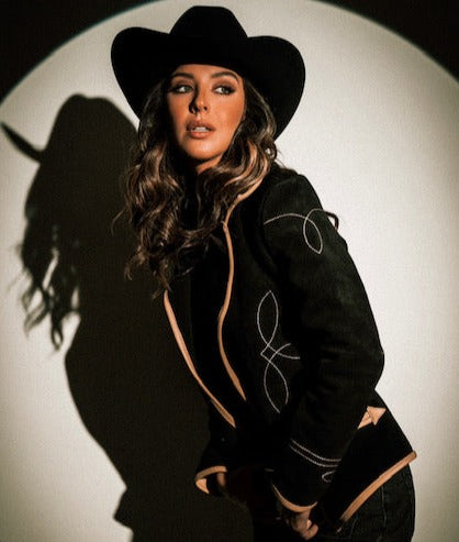 A woman wearing a black suede blazer with tan leather details paired with a black hat