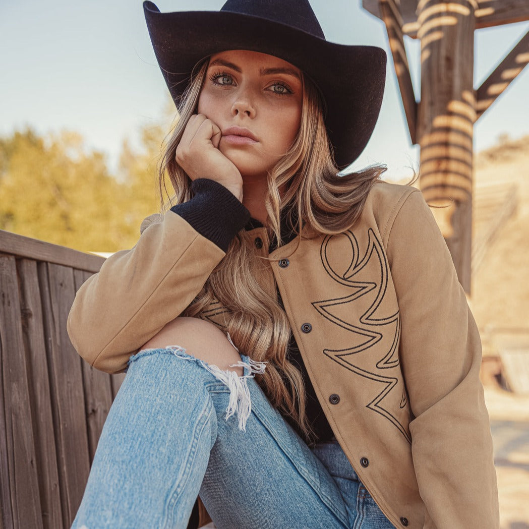 A woman wearing a beige bomber jacket with black western embroidery,  and a wide-brimmed hat.
