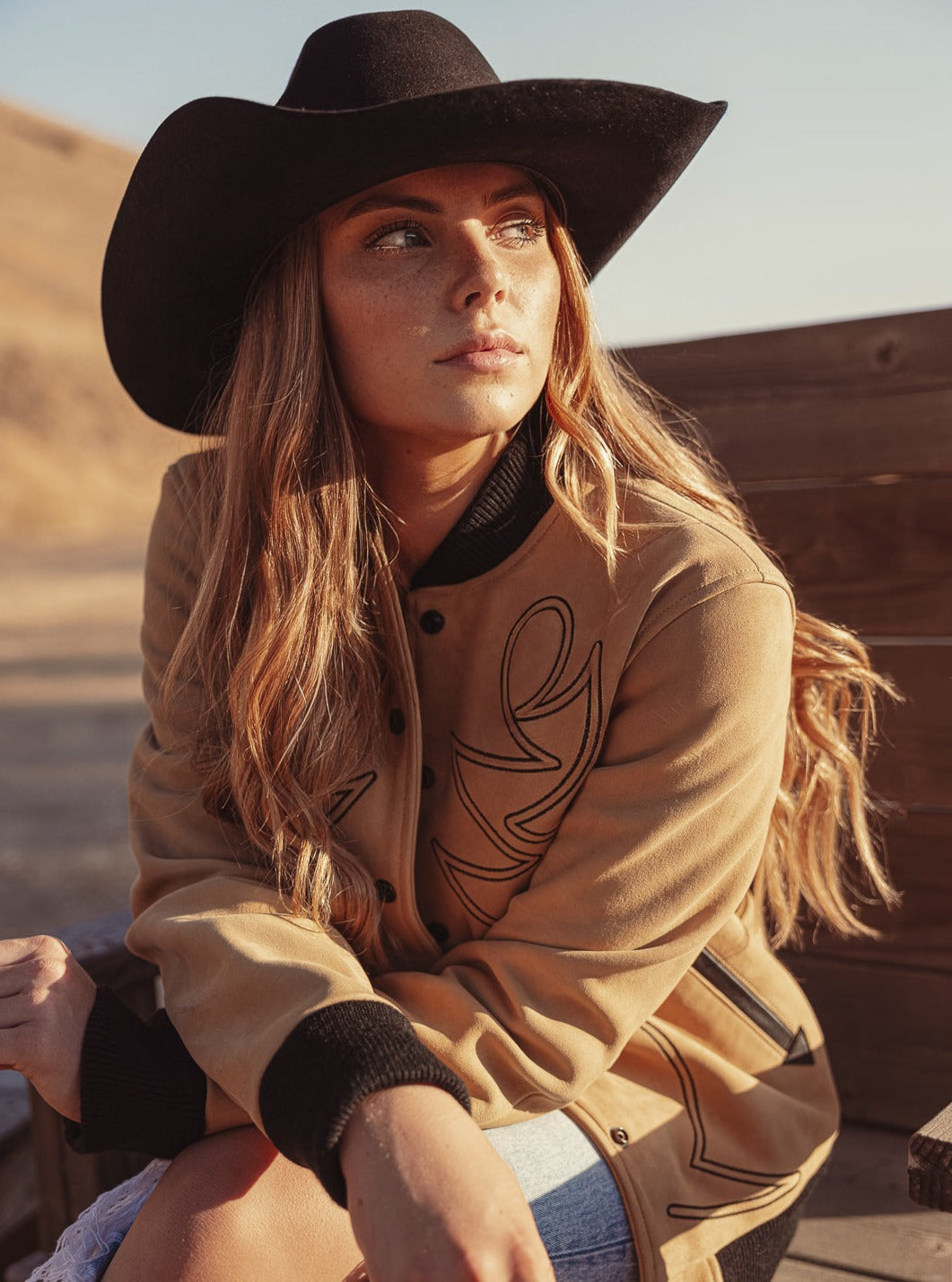 A woman wearing a beige bomber jacket with black western embroidery, and a wide-brimmed cowboy hat