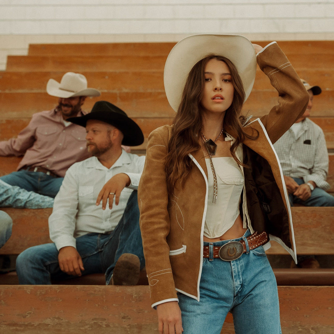 A woman sitting on wooden bleachers wearing a brown carmel suede blazer with white leather piping and details, paired with blue jeans.