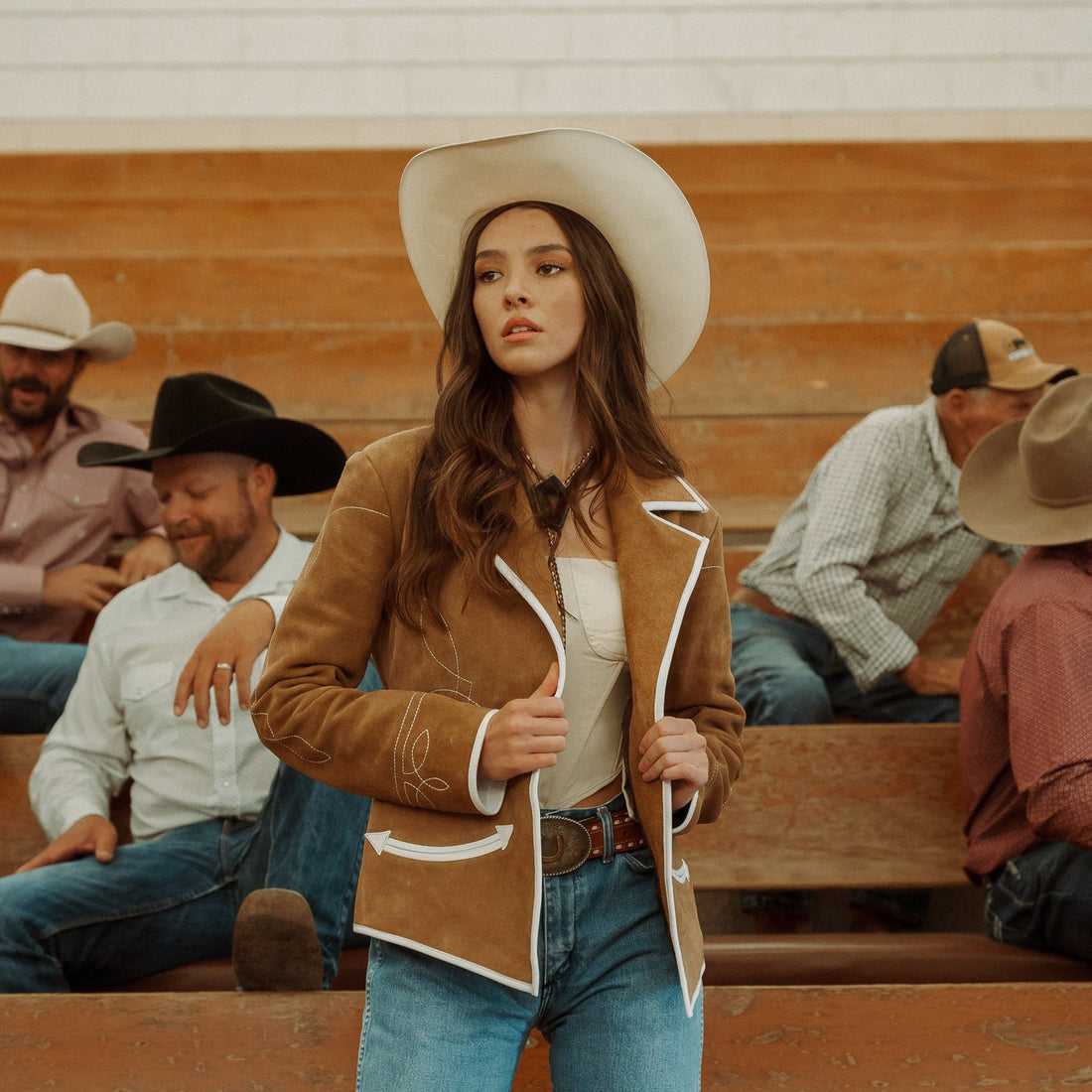A woman sitting on wooden bleachers wearing a brown carmel suede blazer with white leather piping and details, paired with blue jeans.