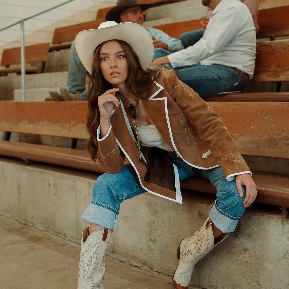 A woman sitting on wooden bleachers wearing a brown carmel suede blazer with white leather piping and details, paired with blue jeans.
