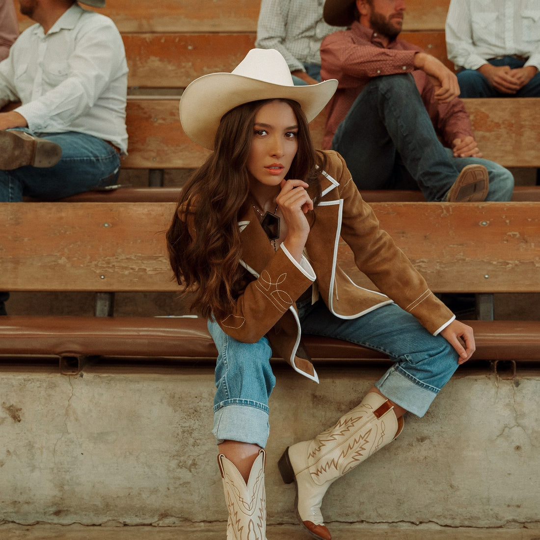 A woman sitting on wooden bleachers wearing a brown carmel suede blazer with white leather piping and details, paired with blue jeans.