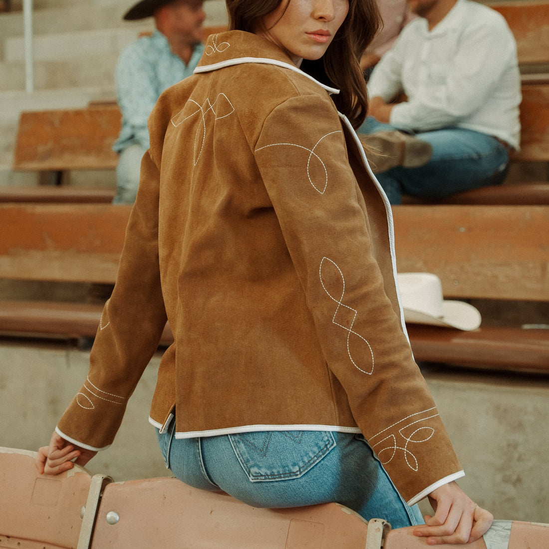 A woman sitting on wooden bleachers wearing a brown carmel suede blazer with white leather piping and details, paired with blue jeans.