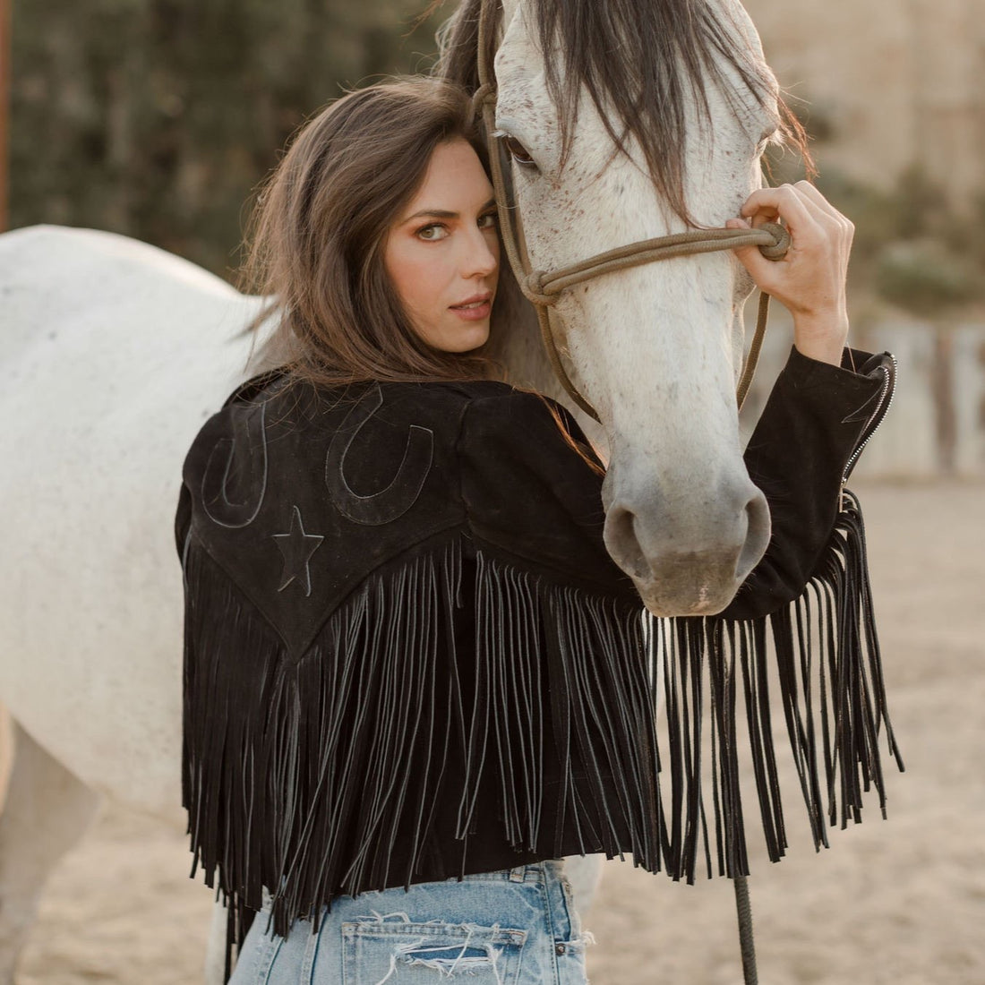 A woman standing next to a white horse, wearing a black suede jacket with fringe on the arms horseshoe embroidered on the back, paired with blue jeans.