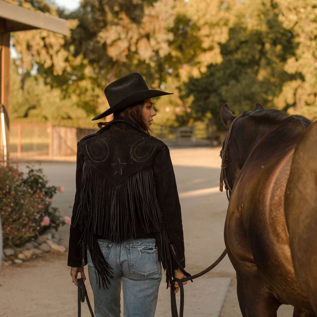 A woman standing next to a horse, wearing a black suede jacket with fringe on the arms horseshoe embroidered on the back, paired with blue jeans.