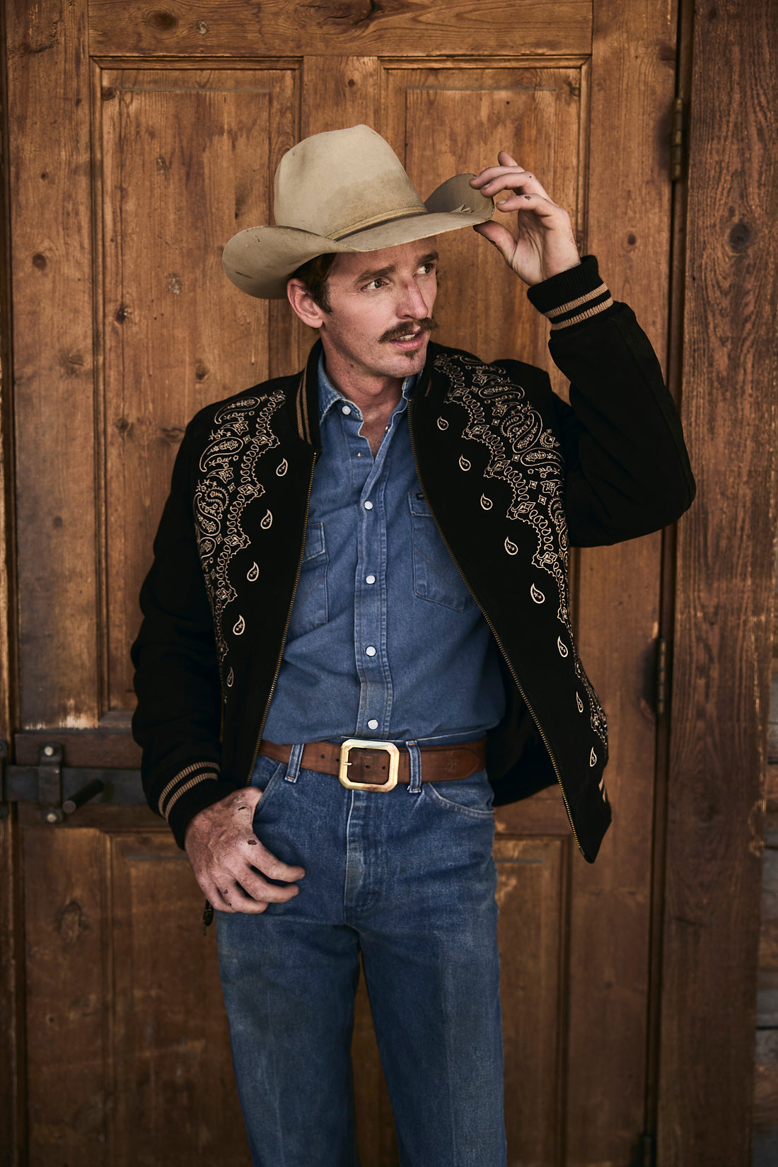 Man in cowboy hat and suede bandana bomber jacket by Western & Co leaning  against a wooden wall 