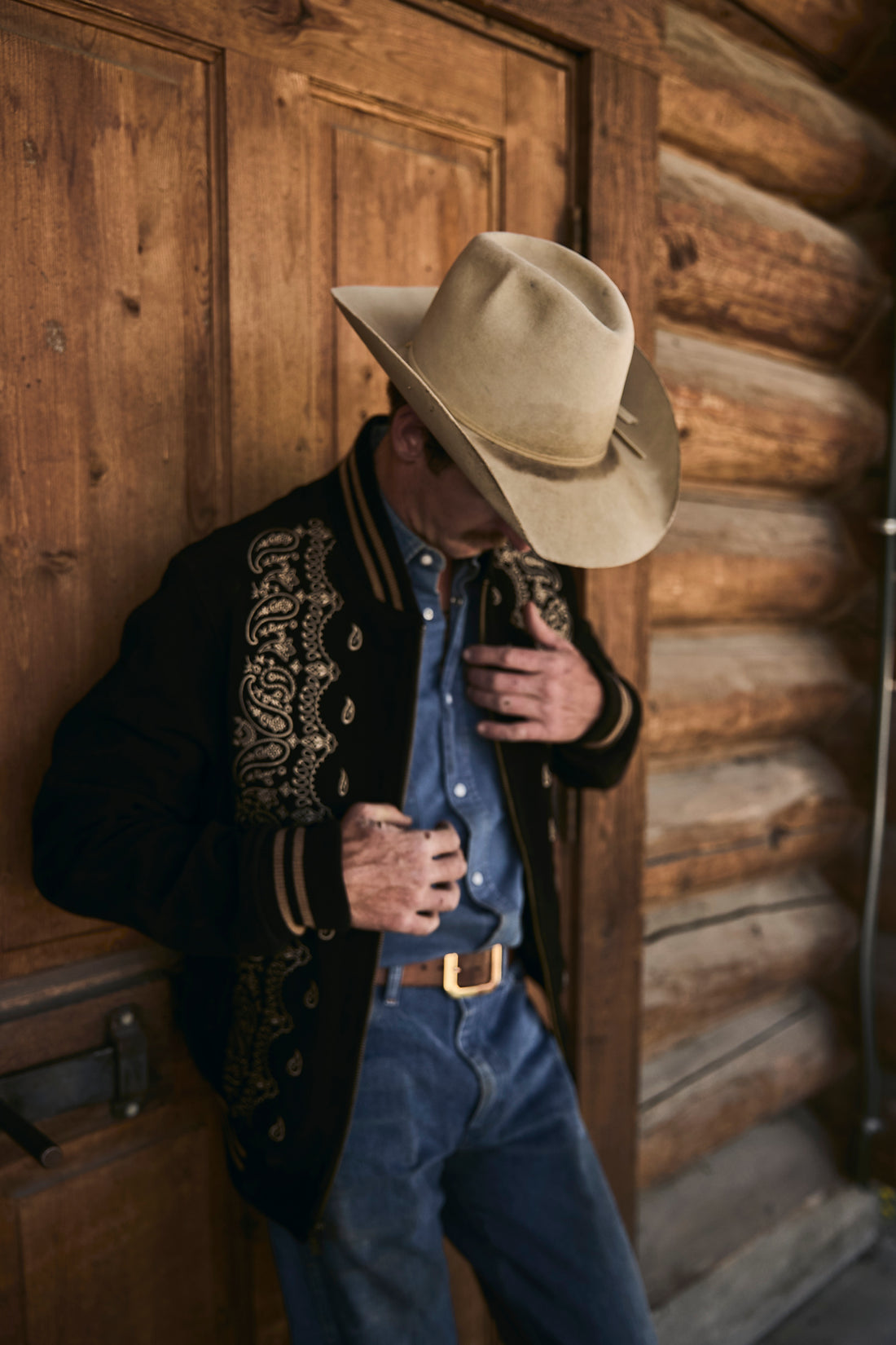 Man in cowboy hat and suede bandana bomber jacket by Western & Co leaning  against a wooden wall 