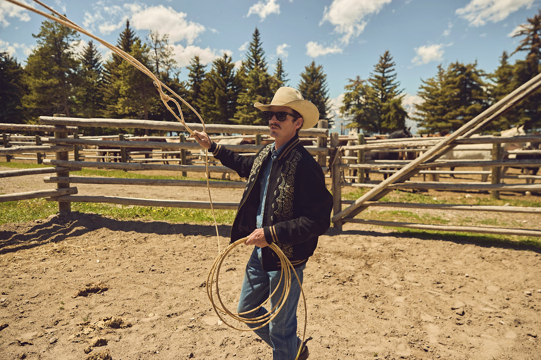 Man in cowboy hat and suede bandana bomber jacket by Western & Co holding a lasso in a rural setting with wooden fences and trees