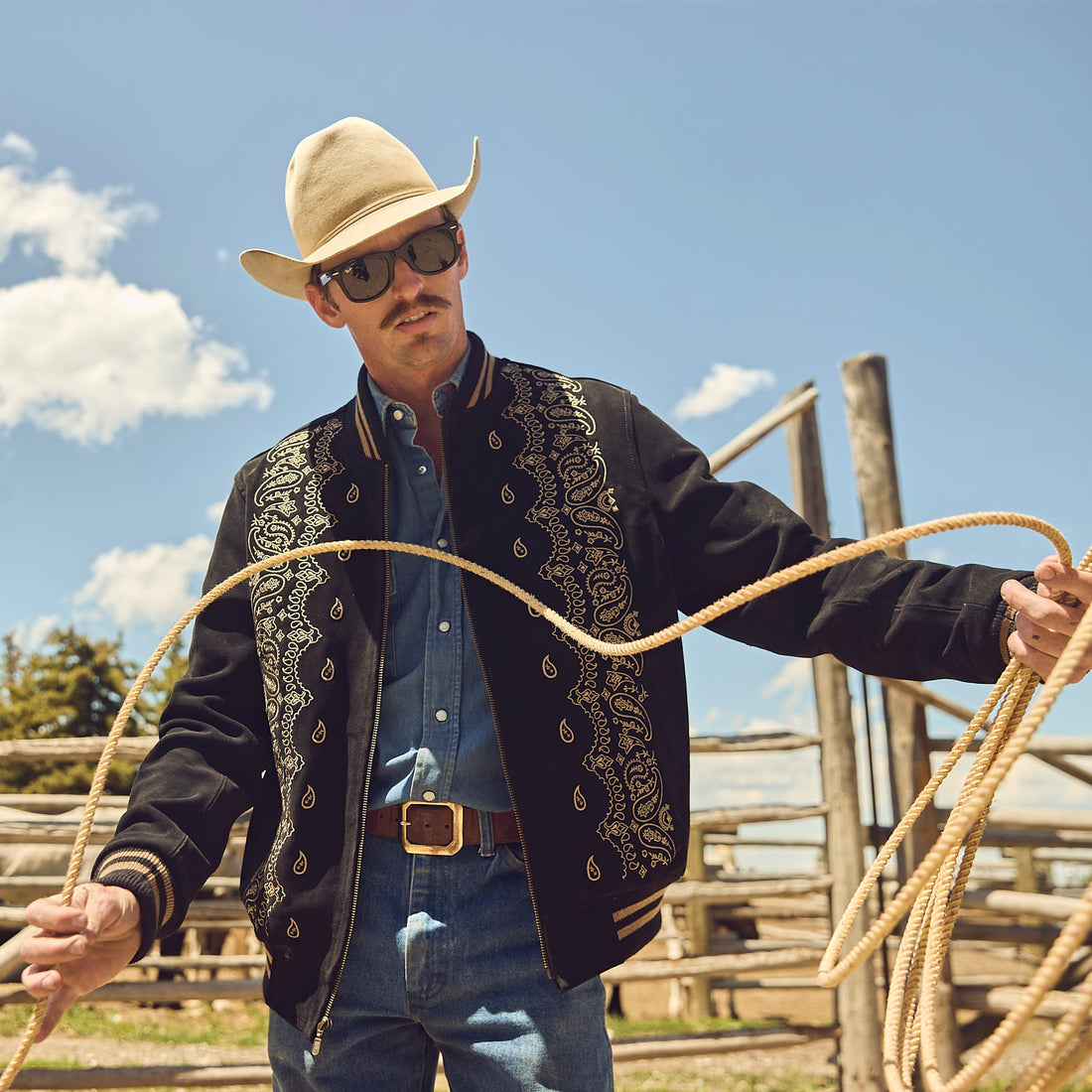 Man in cowboy hat and suede bandana bomber jacket holding a rope with a fence and sky in the background
