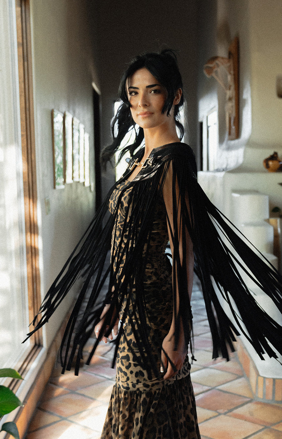 A woman wearing a leopard-print dress with dramatic black fringe draped over her shoulders, standing in a sunlit indoor hallway with tiled floors and wall decor.