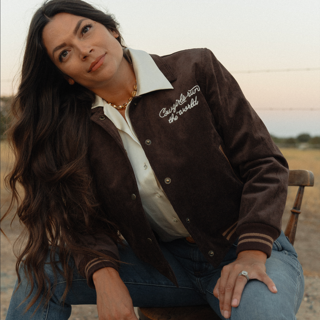 A woman wearing a dark brown suede varsity-style jacket with white script embroidery on the chest, layered over a cream button-up shirt and blue jeans, sitting on a wooden chair in an outdoor desert setting.