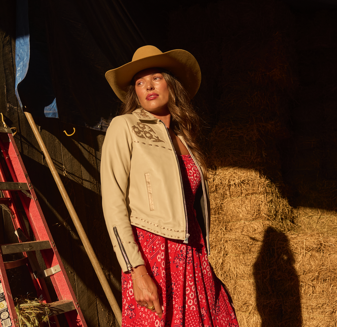 Woman in cowboy hat, red dress and a white embroidered jacket by Western & Co next to ladder and hay bales