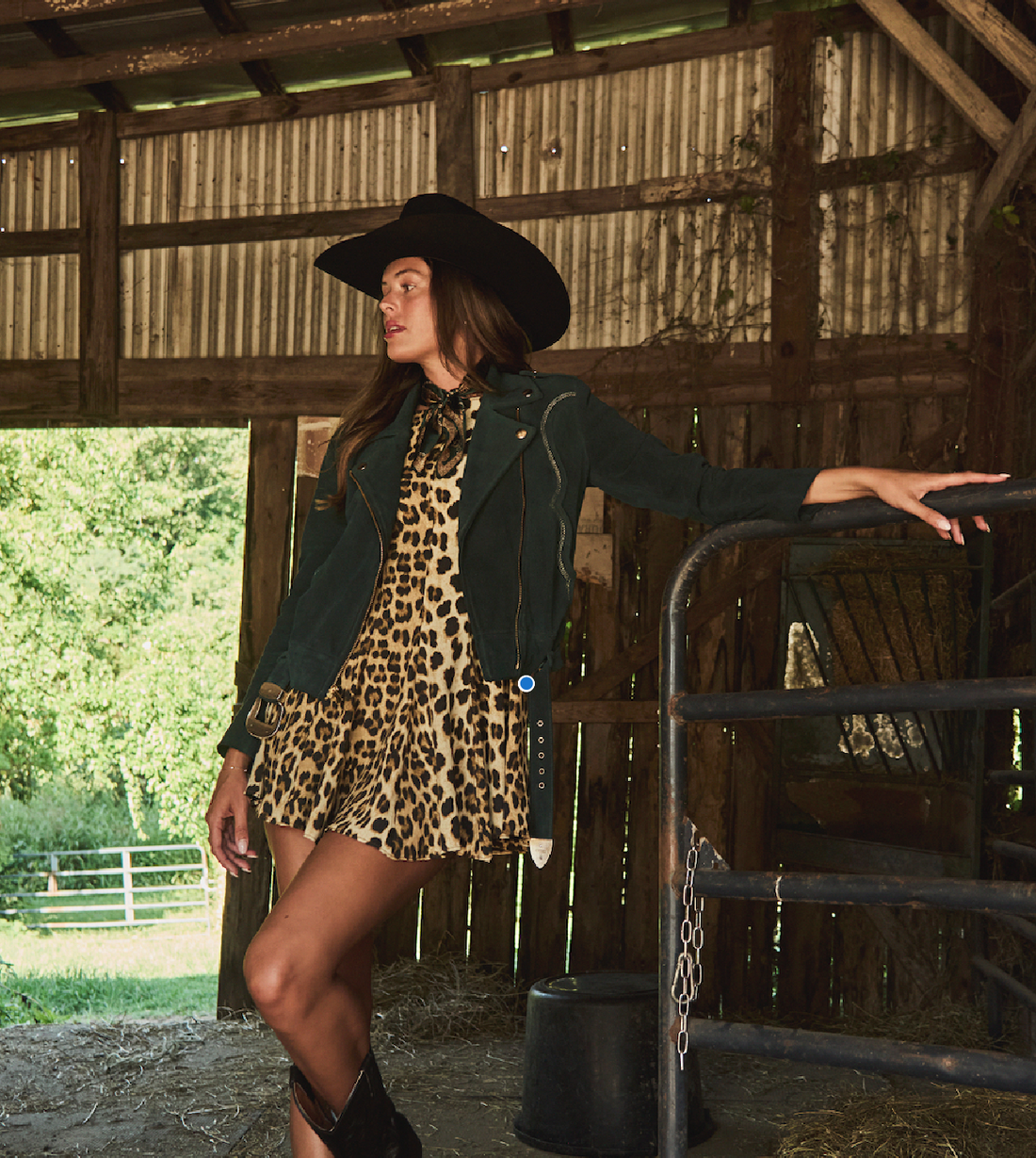 Woman in a cowboy hat and leopard dress wearing emerald green biker jacket by Western & Co standing in a barn 