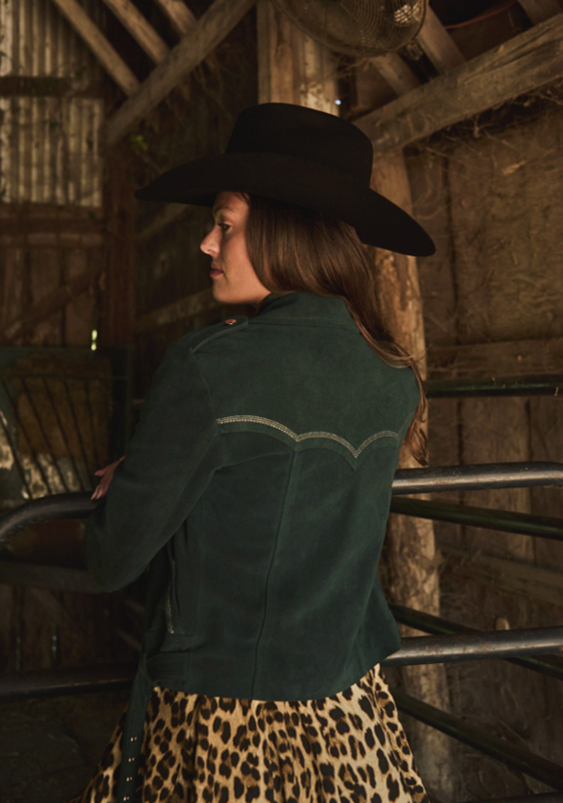 Woman in a cowboy hat and leopard dress wearing emerald green biker jacket by Western & Co standing in a barn setting 
