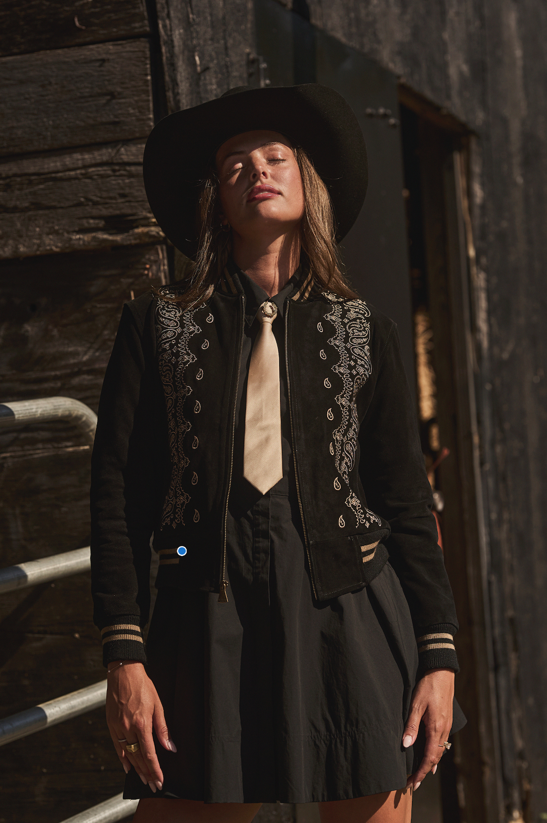 Woman in a cowboy hat wearing black embroidered bomber jacket by Western & Co against a wooden wall 
