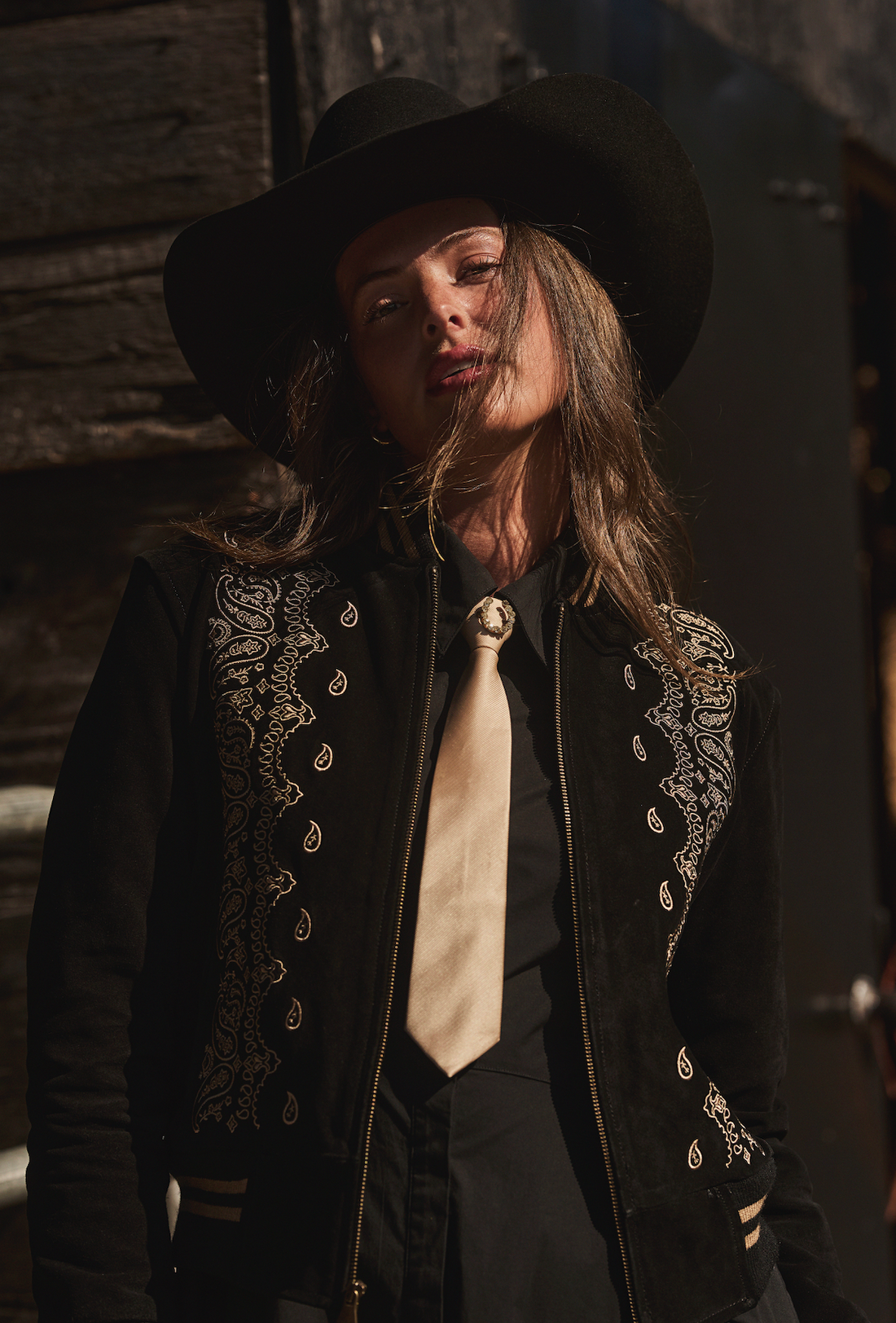 Woman in a cowboy hat wearing black embroidered bomber jacket by Western & Co against a wooden wall 
