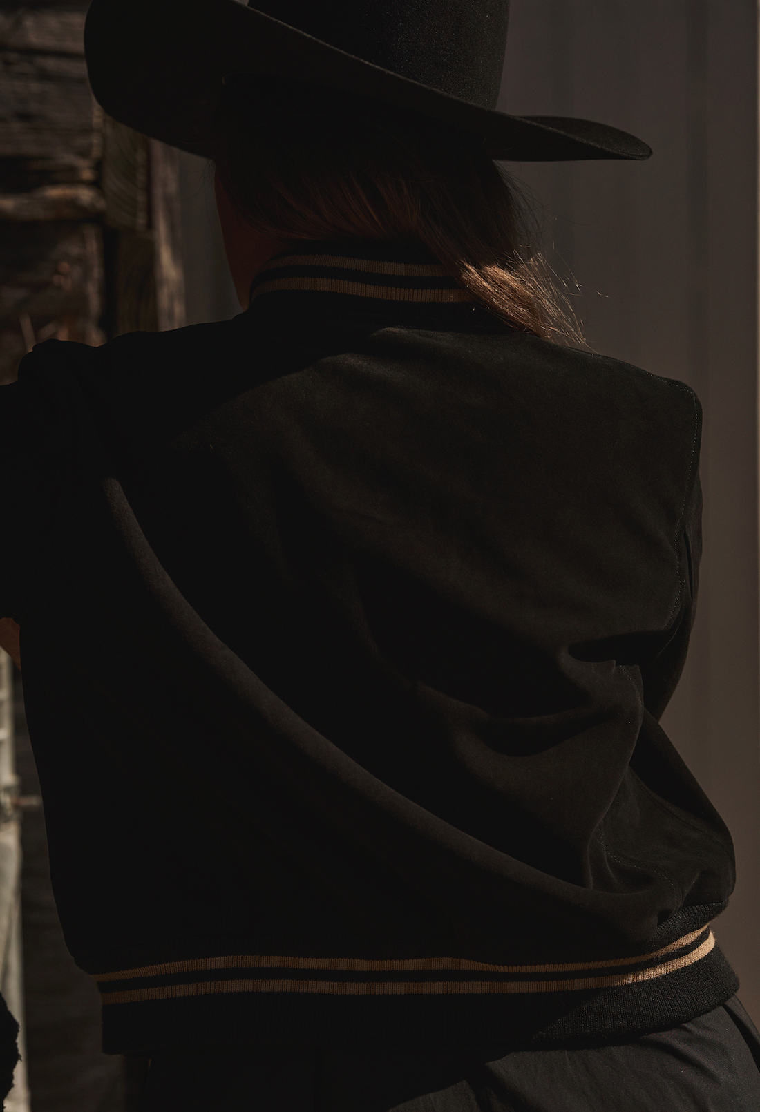 Woman in a cowboy hat wearing black embroidered bomber jacket by Western & Co against a wooden wall 