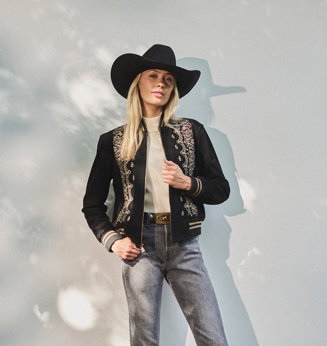 Woman in a cowboy hat wearing black embroidered bomber jacket by Western & Co against a light background 